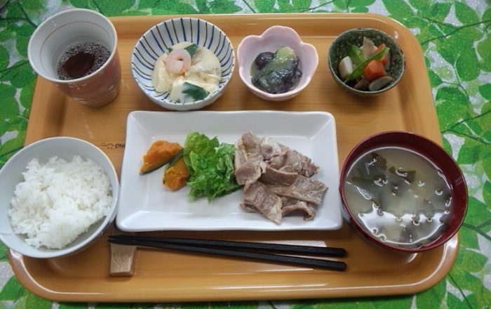 介護施設の食事風景。木製トレイに白いご飯、肉料理、小鉢、汁物が配膳された栄養バランスの取れた食事。