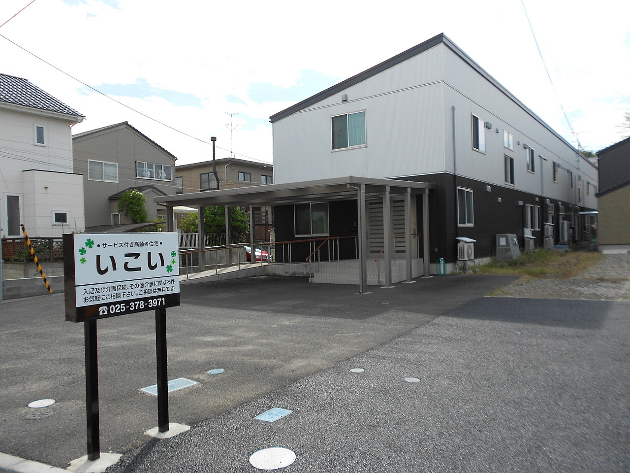 介護施設「いこい」の建物外観。白と黒の配色の2階建て施設で、玄関前に看板が設置されている。